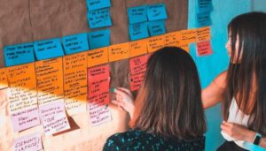 Duas mulheres organizando post-its coloridos em uma parede, representando os passos para planejar um cronograma de postagens no Instagram.