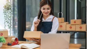 Foto de uma mulher segurando uma caixa de produtos e olhando para um laptop, representando táticas comprovadas para vender pelo Instagram.