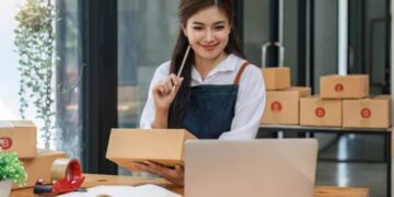 Foto de uma mulher segurando uma caixa de produtos e olhando para um laptop, representando táticas comprovadas para vender pelo Instagram.