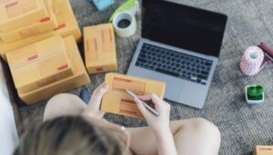 Foto de uma pessoa preenchendo etiquetas de envio em caixas de produtos ao lado de um laptop, representando a criação de uma loja no Instagram Shopping para vender mais em 2024.