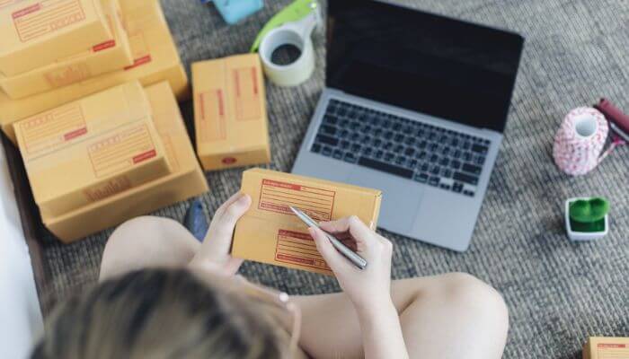 Foto de uma pessoa preenchendo etiquetas de envio em caixas de produtos ao lado de um laptop, representando a criação de uma loja no Instagram Shopping para vender mais em 2024.