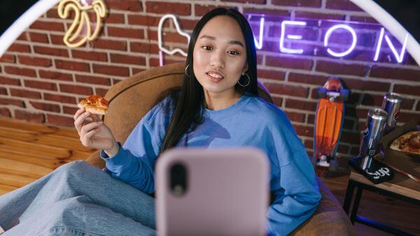 Jovem comendo pizza enquanto grava um vídeo com o celular, cenário com luzes neon ao fundo.