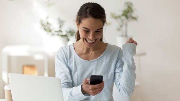 Mulher sorrindo de forma entusiasmada enquanto segura o celular e senta-se em frente ao computador.