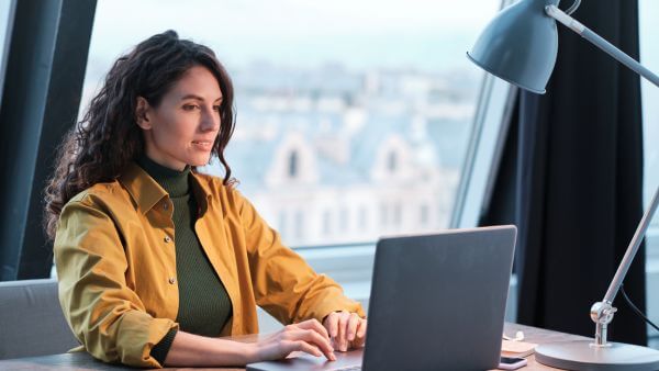 Mulher com cabelo cacheado, sentada em um escritório moderno, usando um notebook e procurando por legenda photo dump. Ao fundo, uma vista para prédios. Contexto de produtividade e tecnologia.